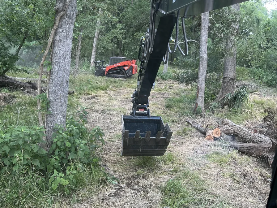 Excavator bucket among cut logs with a mulching skid steer behind in the woods, land clearing